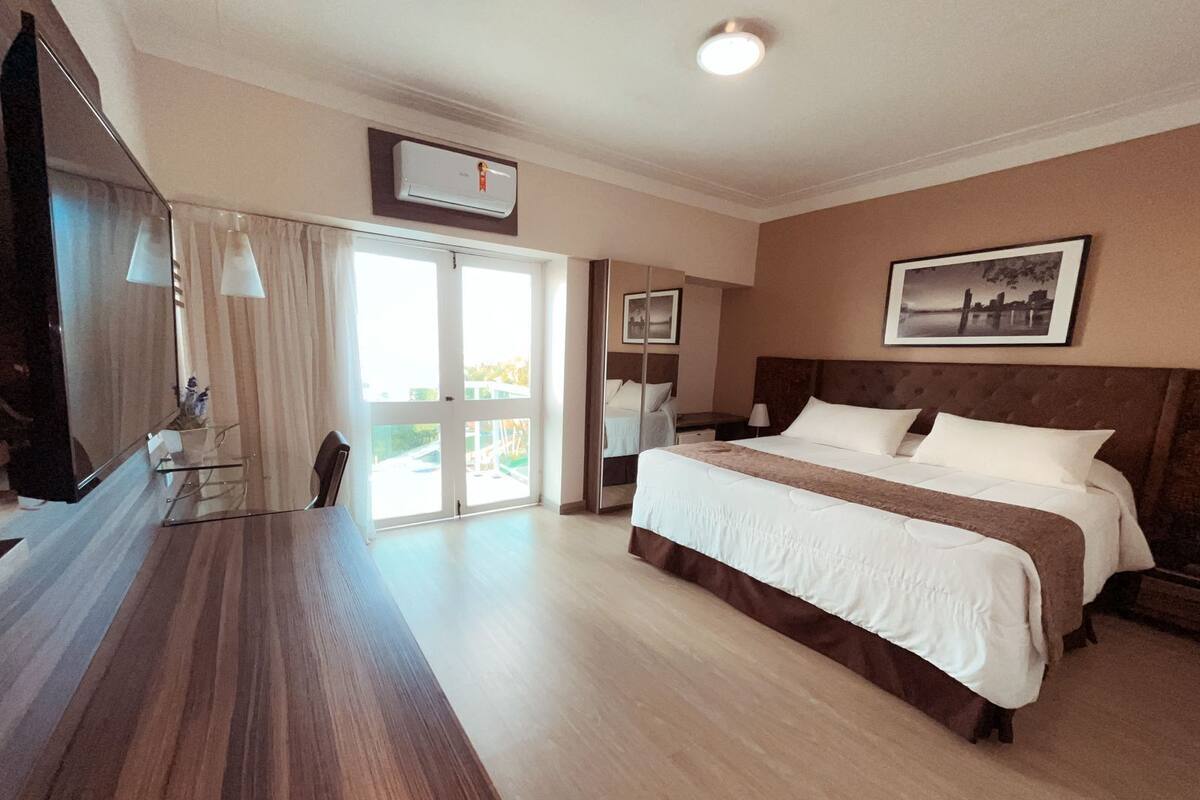 Une chambre d’hôtel avec un grand lit, un bureau, une chaise, une télévision et une fenêtre avec des rideaux.
