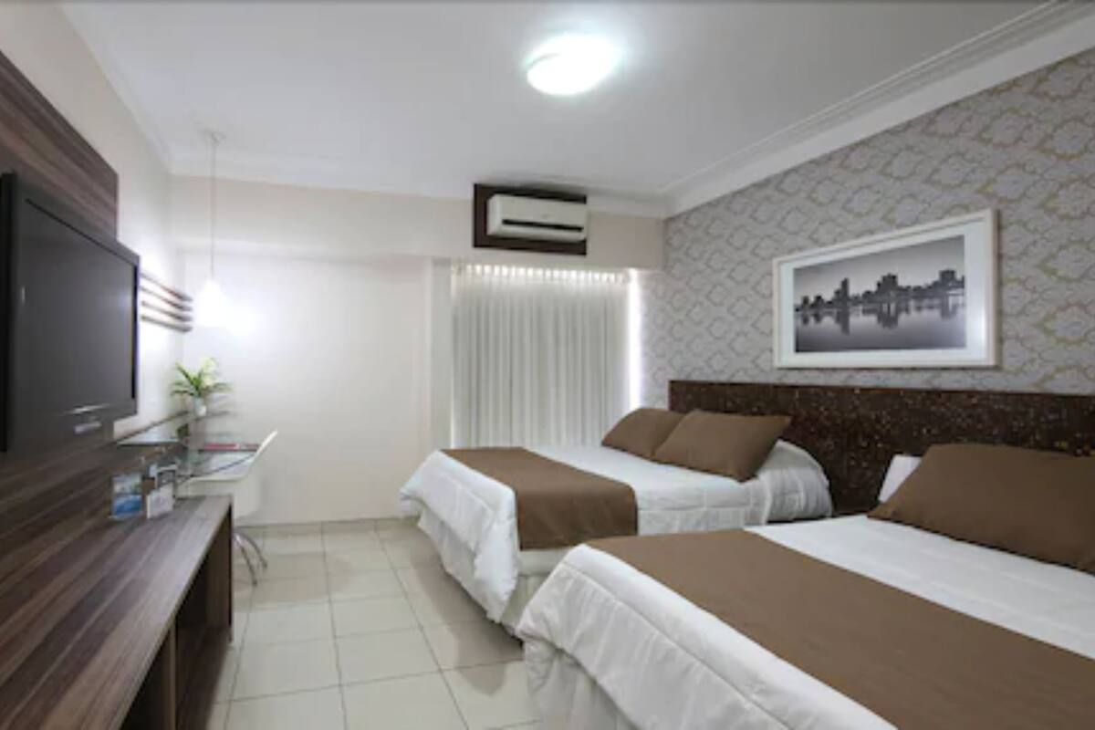 Une chambre d’hôtel avec deux lits, une télévision, un bureau agrémenté d’une plante et un tableau représentant un paysage urbain accroché au mur.