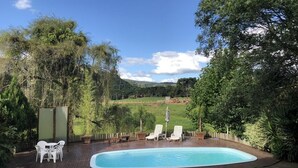 Une piscine entourée d’une terrasse en bois et de verdure, avec des chaises longues et une table à proximité.