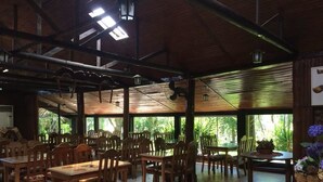 Un vaste espace de restauration intérieur, doté de nombreuses tables et chaises en bois, de grandes fenêtres laissant entrer la lumière naturelle et d’un plafond avec des poutres apparentes et des luminaires suspendus.