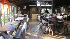 Une terrasse extérieure en bois, dotée de tables et de bancs, et entourée de fenêtres ornées de rideaux.