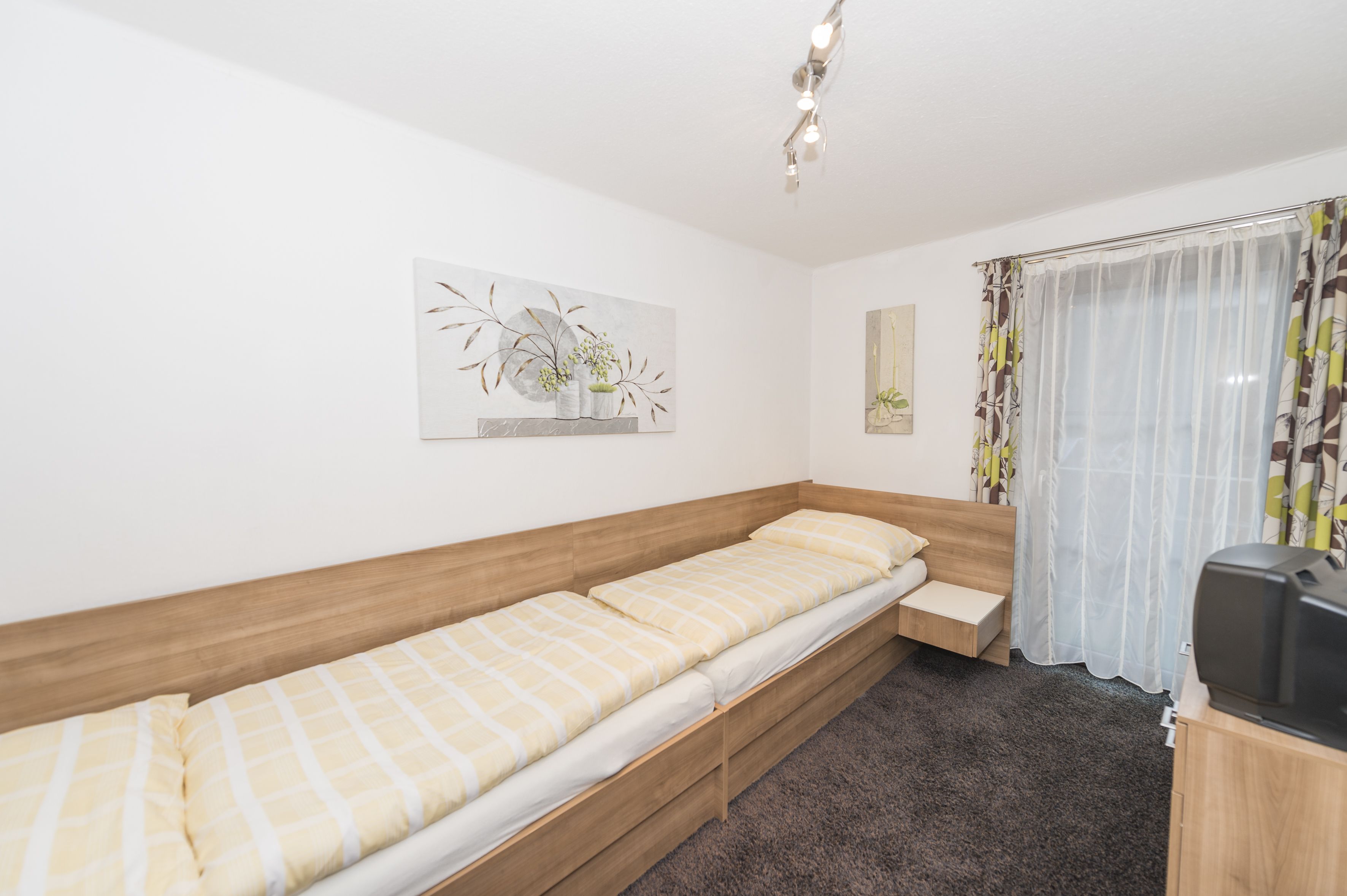 Una cama individual con cabecera de madera, una mesita de noche pequeña y un televisor sobre un mueble de madera.