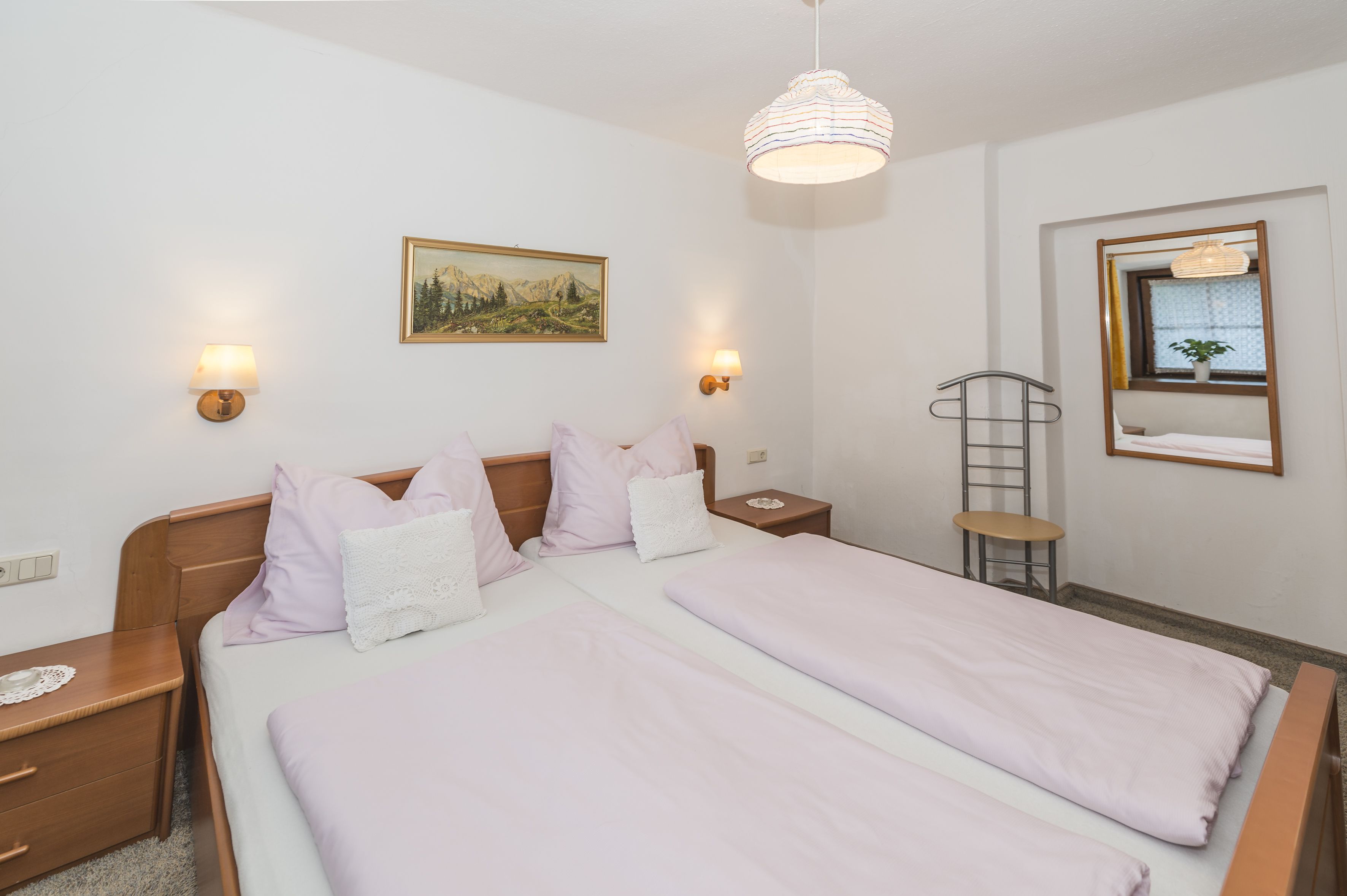 A double bed with a wooden headboard, two bedside tables, a wall-mounted lamp, a small table with a chair, a ladder, and a framed picture on the wall.
