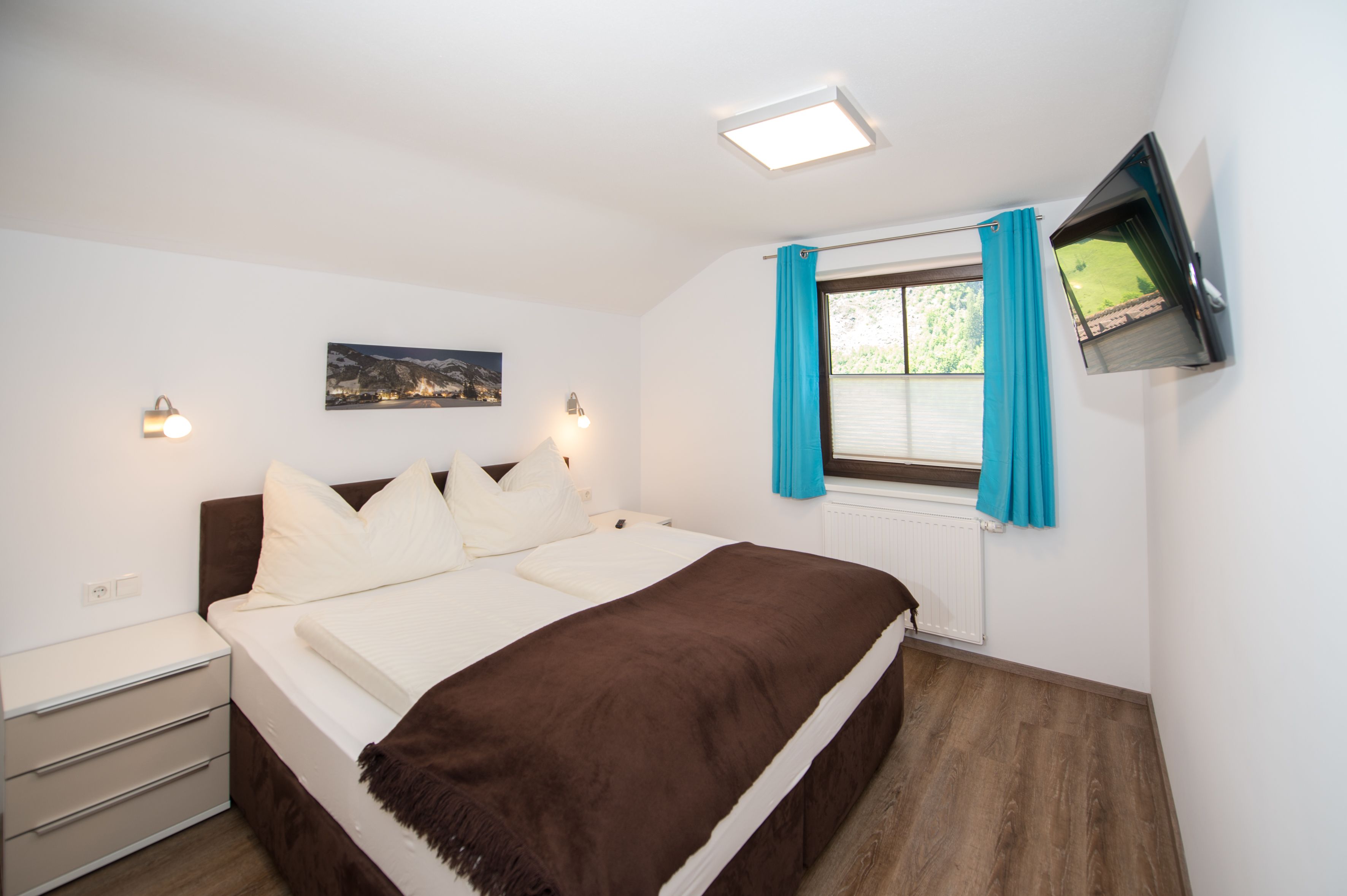 Un dormitorio con cama, mesita de noche, televisor en la pared y ventana con cortinas.