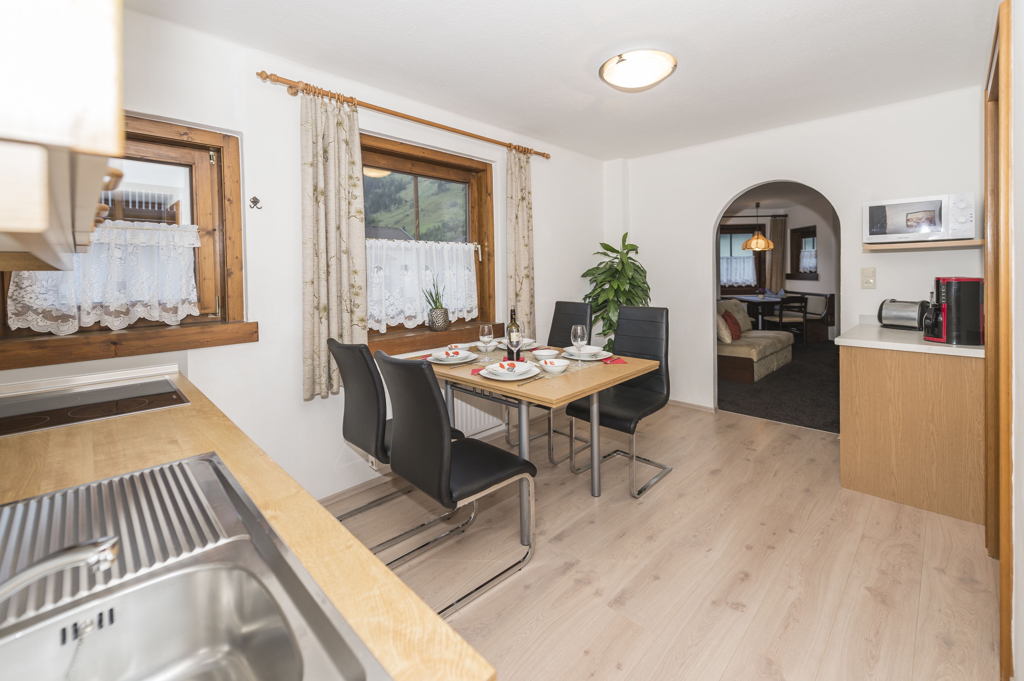 A modern kitchen with a sink, dining table, and chairs. There is a window with curtains, a potted plant, and a microwave on a shelf.