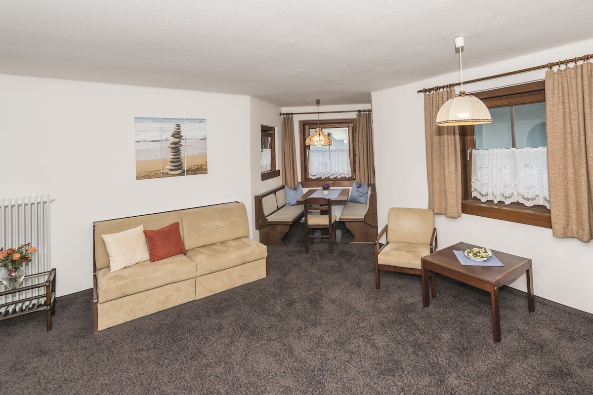 A furnished room with a sofa, a small table, a chair, a dining table with chairs, a window with curtains, and a hanging light fixture.