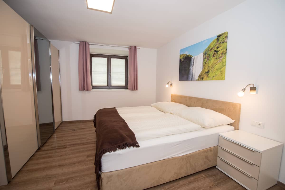 A bedroom with a bed, a nightstand, a window with curtains, a wall-mounted light, and a painting.