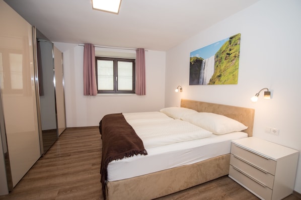A bedroom with a bed, a nightstand, a window with curtains, a wall-mounted light, and a painting.