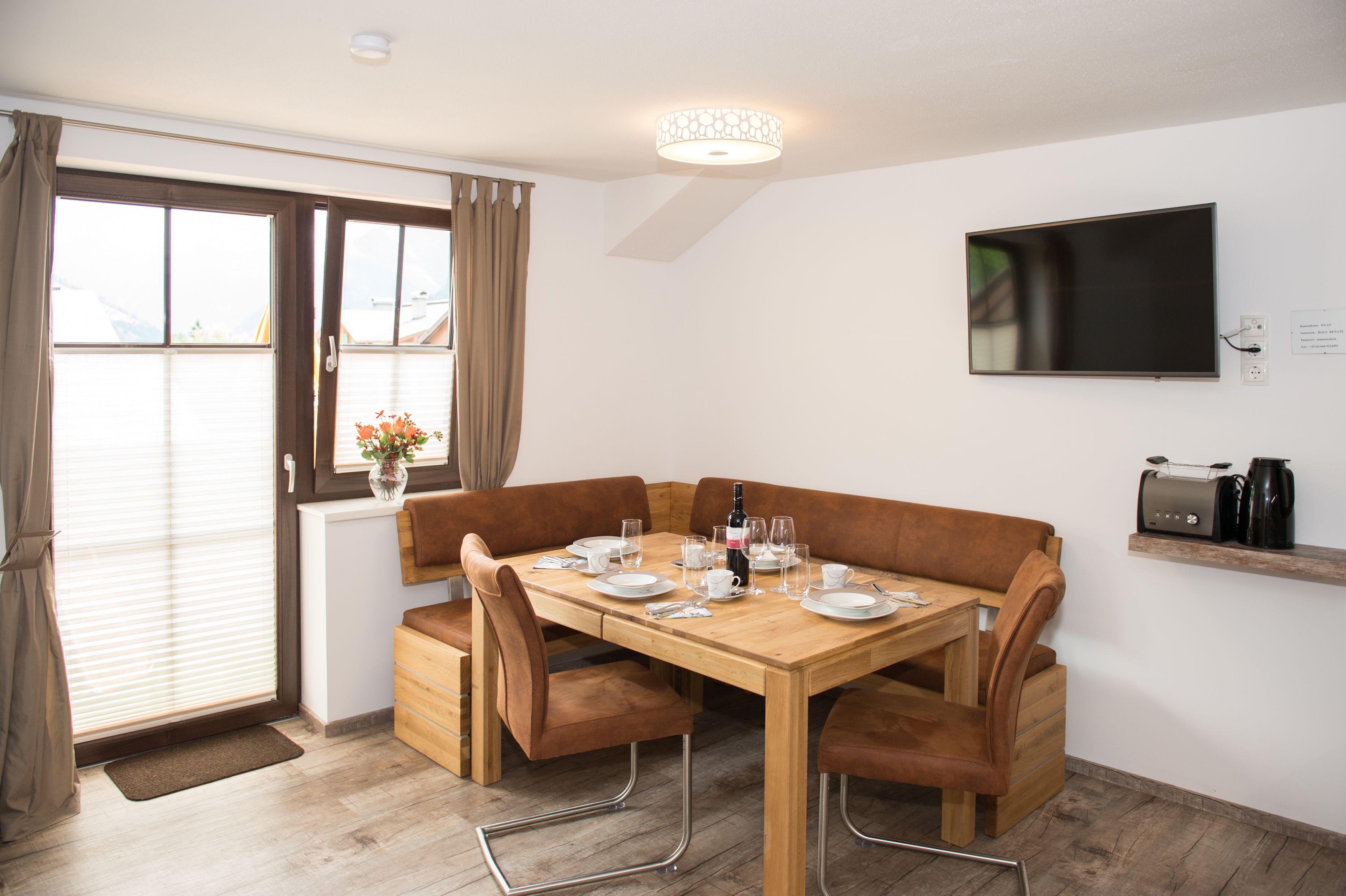 Een eetgedeelte met een houten tafel voor vier personen, beklede banken, een vaas met bloemen en een aan de muur bevestigde televisie.