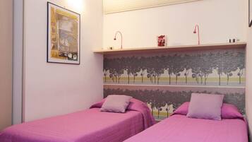 A room with two beds, each with a purple bedspread and pillow, a framed picture on the wall, and a shelf with decorative items.