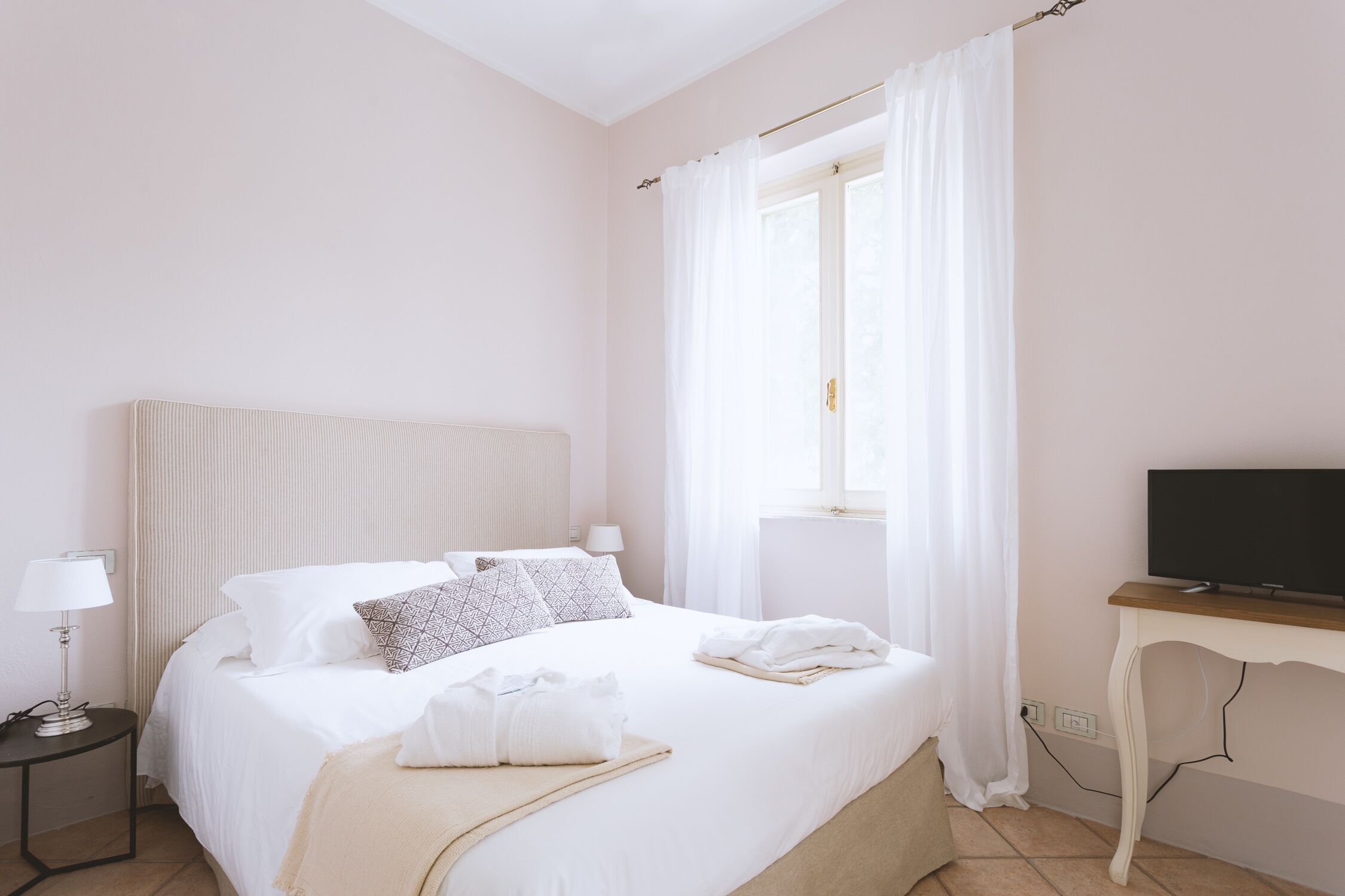 Habitación de hotel ordenada con cama, lámparas de noche, un televisor sobre una mesa de madera y una ventana con cortinas.