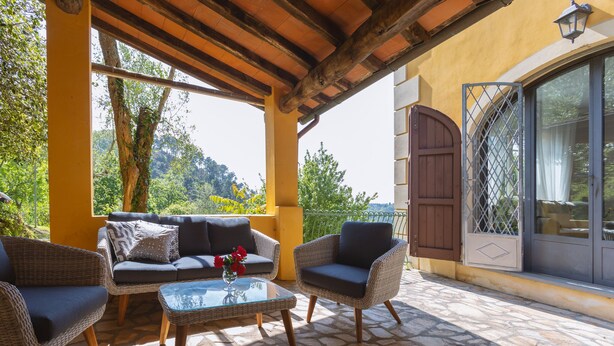 A covered outdoor seating area with wicker furniture, a glass table, and a view of greenery and hills. - Villa Borgovecchio B&B (Camaiore)