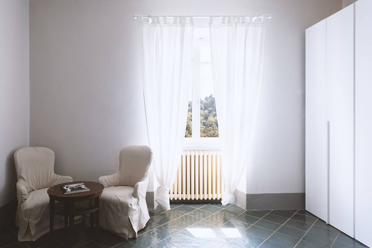 Une pièce avec deux chaises blanches, une petite table ronde, un radiateur et une fenêtre ornée de rideaux légers.