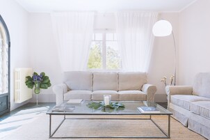 Una sala de estar con un sofá color beige, una mesa de centro de vidrio, una lámpara de pie y una ventana con cortinas translúcidas.