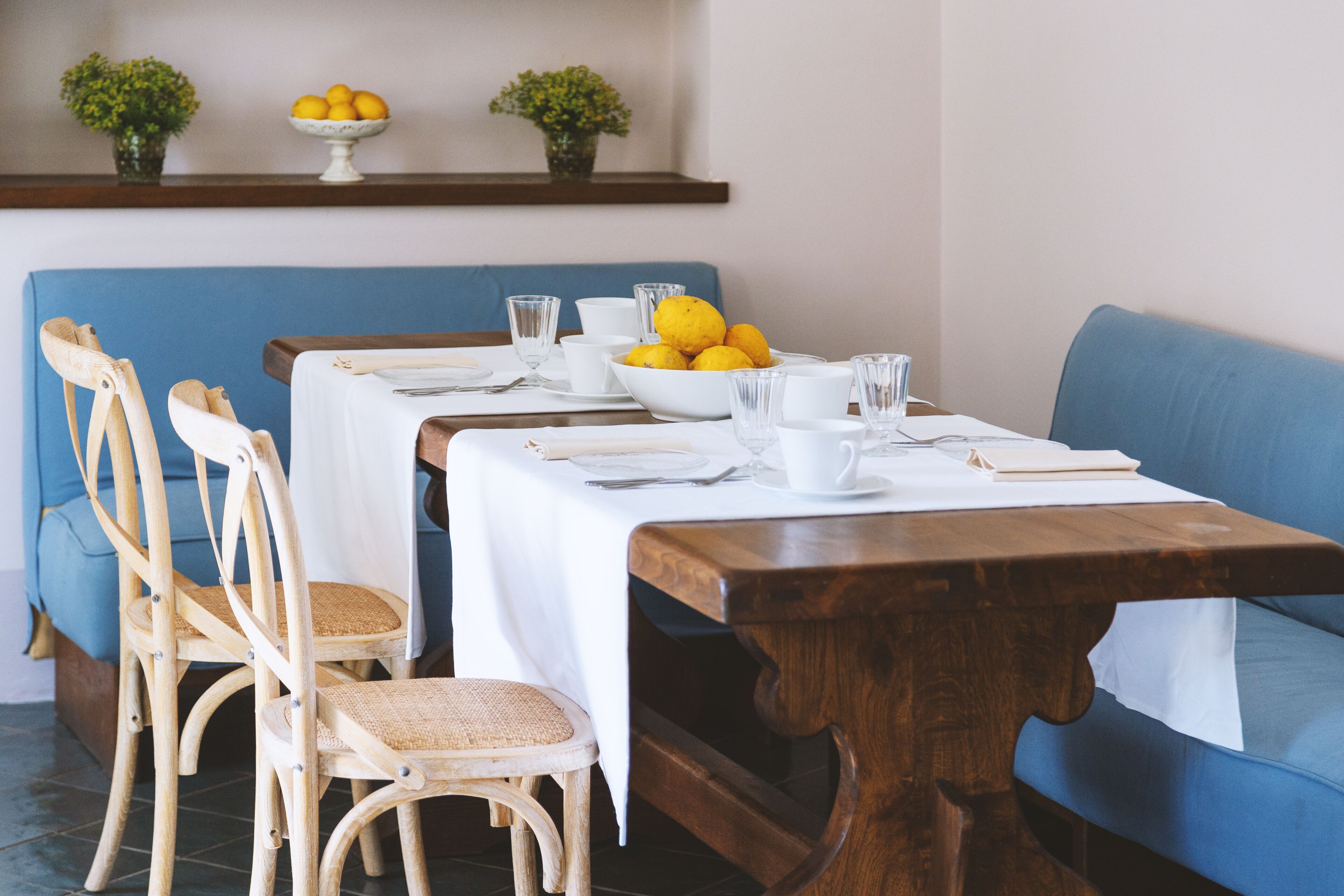 A dining area with a long wooden table set for four, surrounded by wicker chairs. There is a blue sofa in the background and a shelf with a bowl of lemons above it.