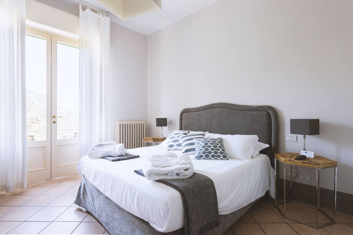 Un lit bien fait, avec une tête de lit grise, du linge de lit blanc et un plaid gris. On y trouve une table de chevet avec une lampe et une petite carte.