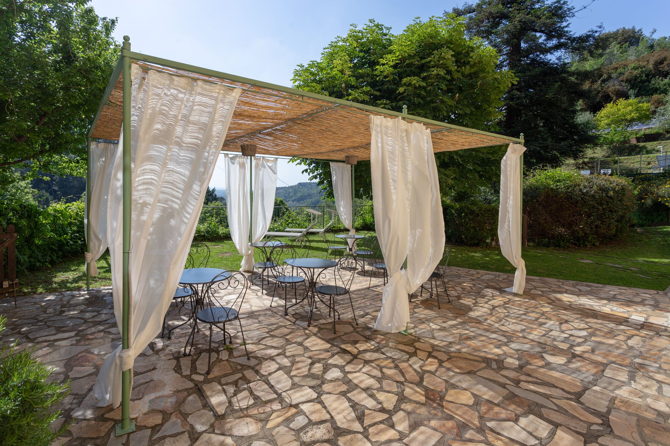 Eine Außenpatio mit Steinboden, überdacht von einer Pergola mit weißen Vorhängen, umgeben von Grünpflanzen und Tischen.