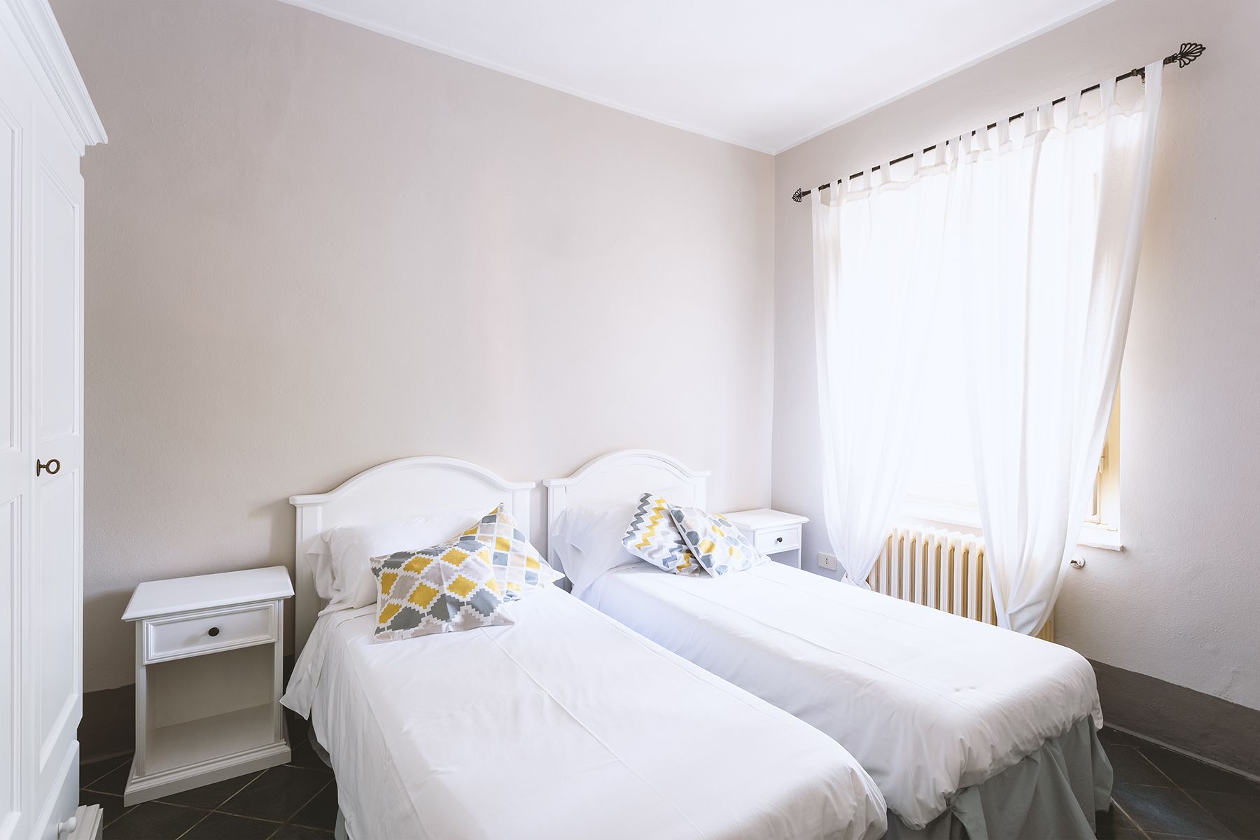 A bedroom with two single beds, a nightstand, a radiator, and a window with curtains.