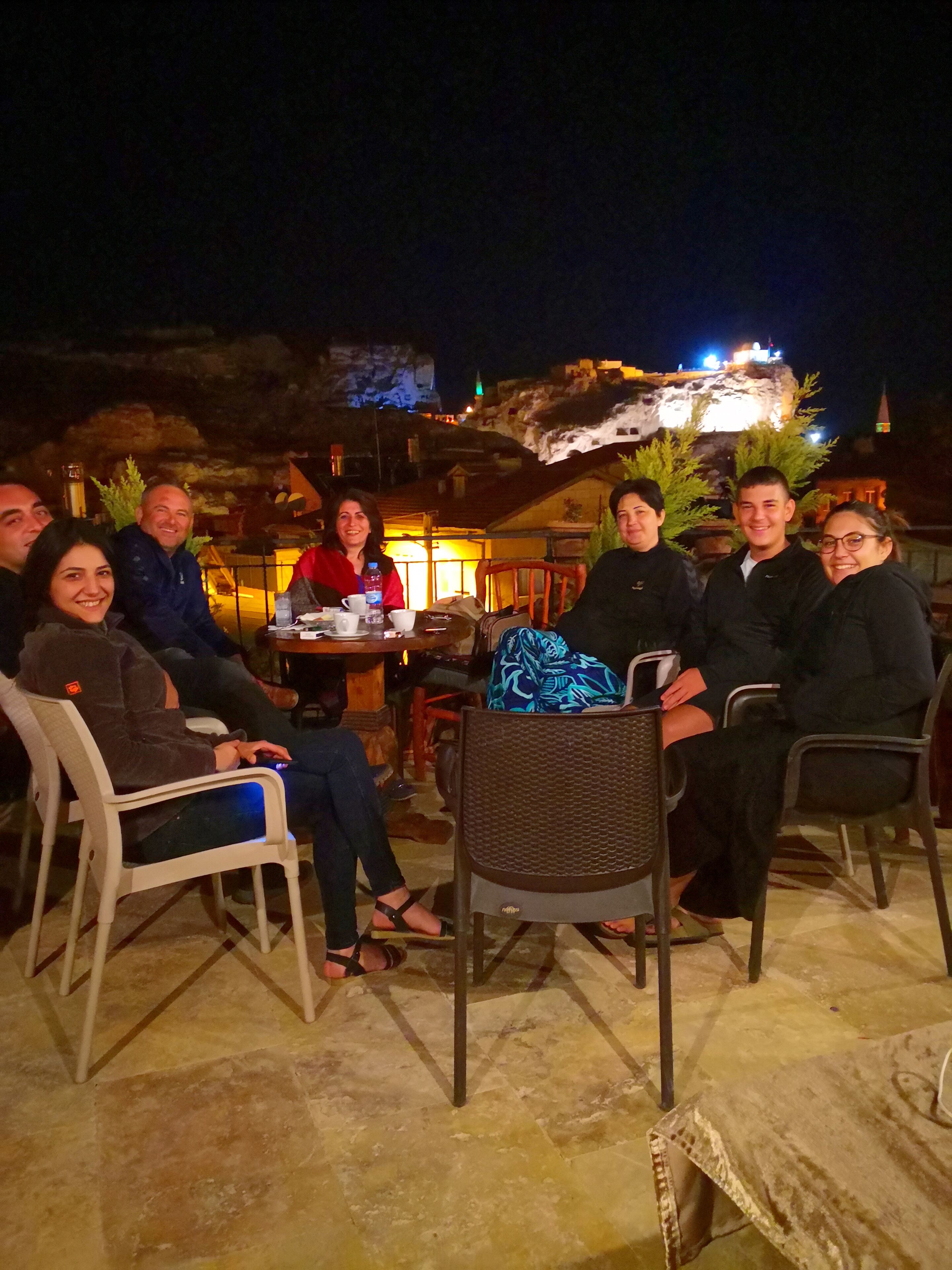 Un grupo de personas sentadas alrededor de una mesa al aire libre por la noche, con un edificio iluminado al fondo.