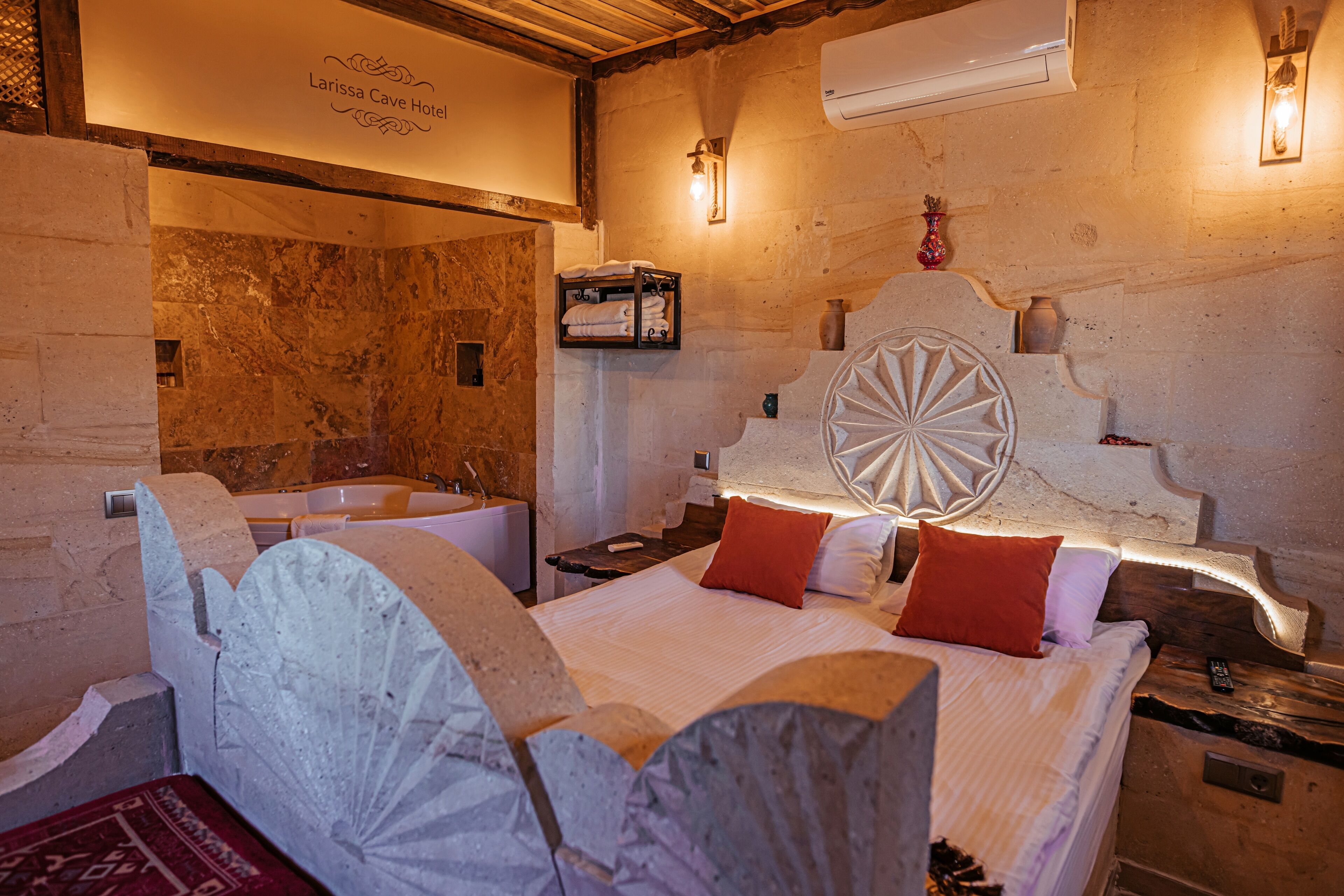 A hotel room with a stone wall, a bed with white bedding and red pillows, a wooden headboard, and a small shelf with towels.