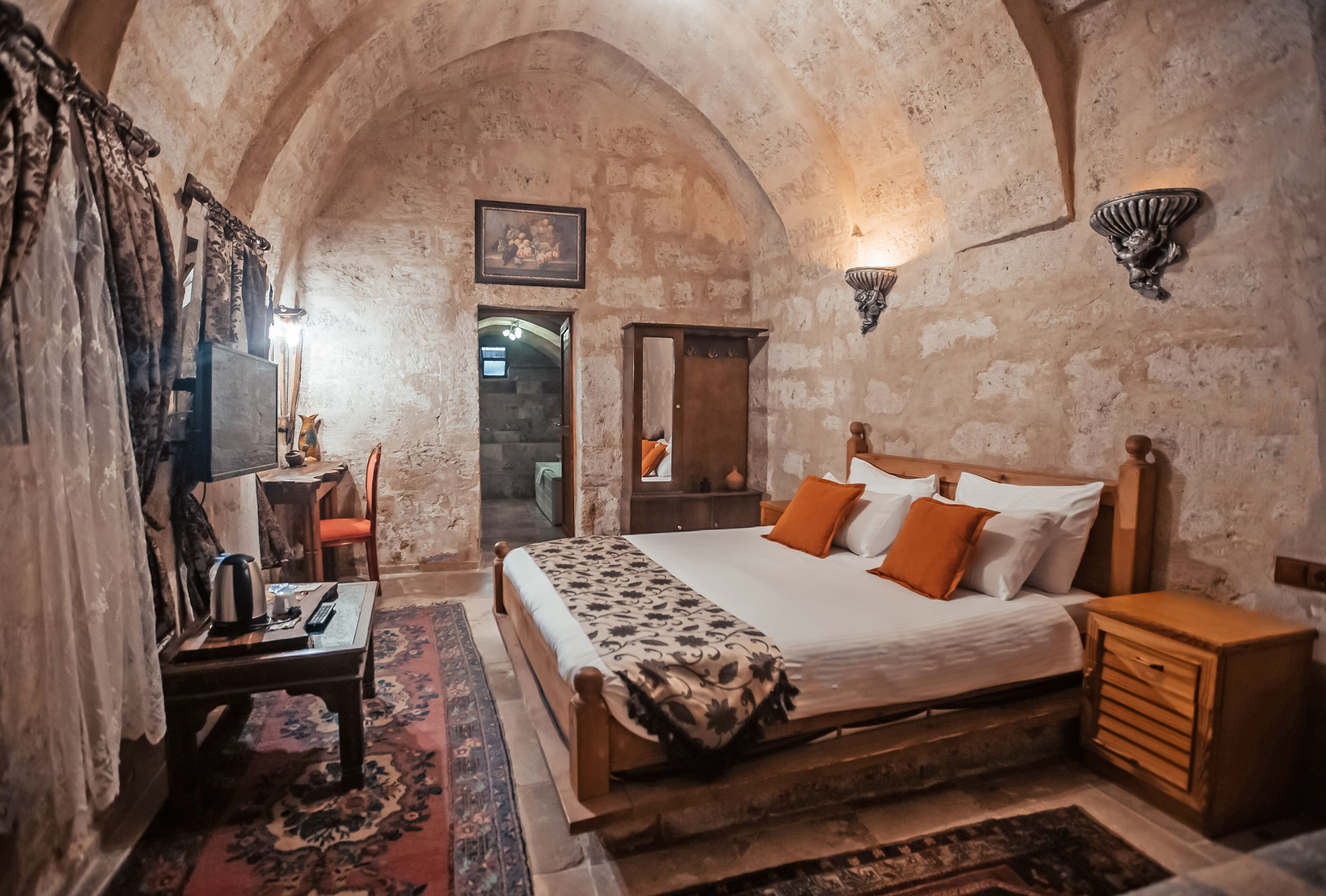 A bedroom with a wooden bed, orange pillows, a nightstand, a mirror, and a stone wall.