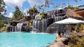 A pool with a waterfall feature, surrounded by tropical vegetation and a clear sky. - Ganso Complexo de Lazer (Lambari)