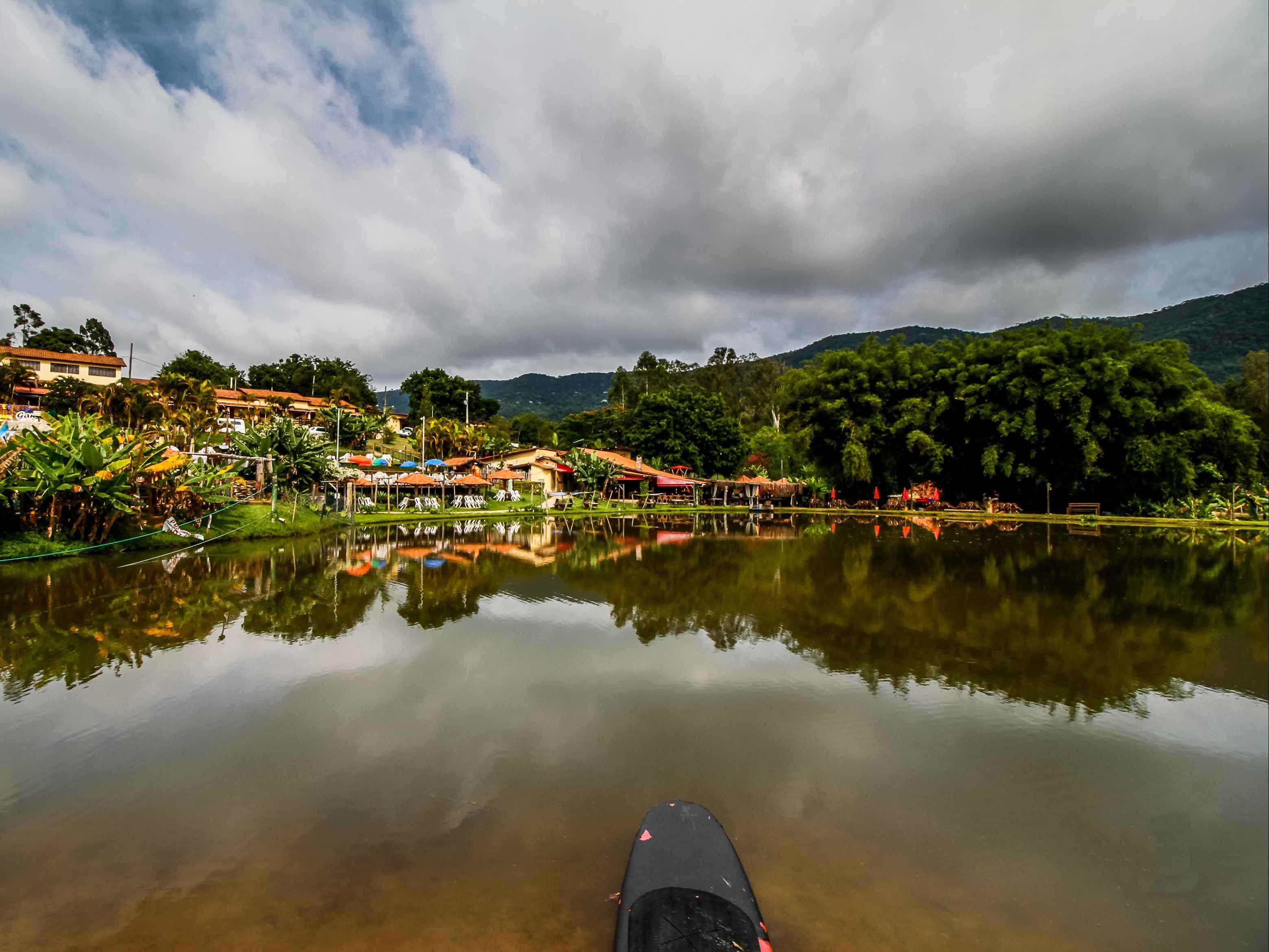 Un lac paisible où se reflètent les bâtiments et les arbres, entouré d’une végétation luxuriante et de collines.