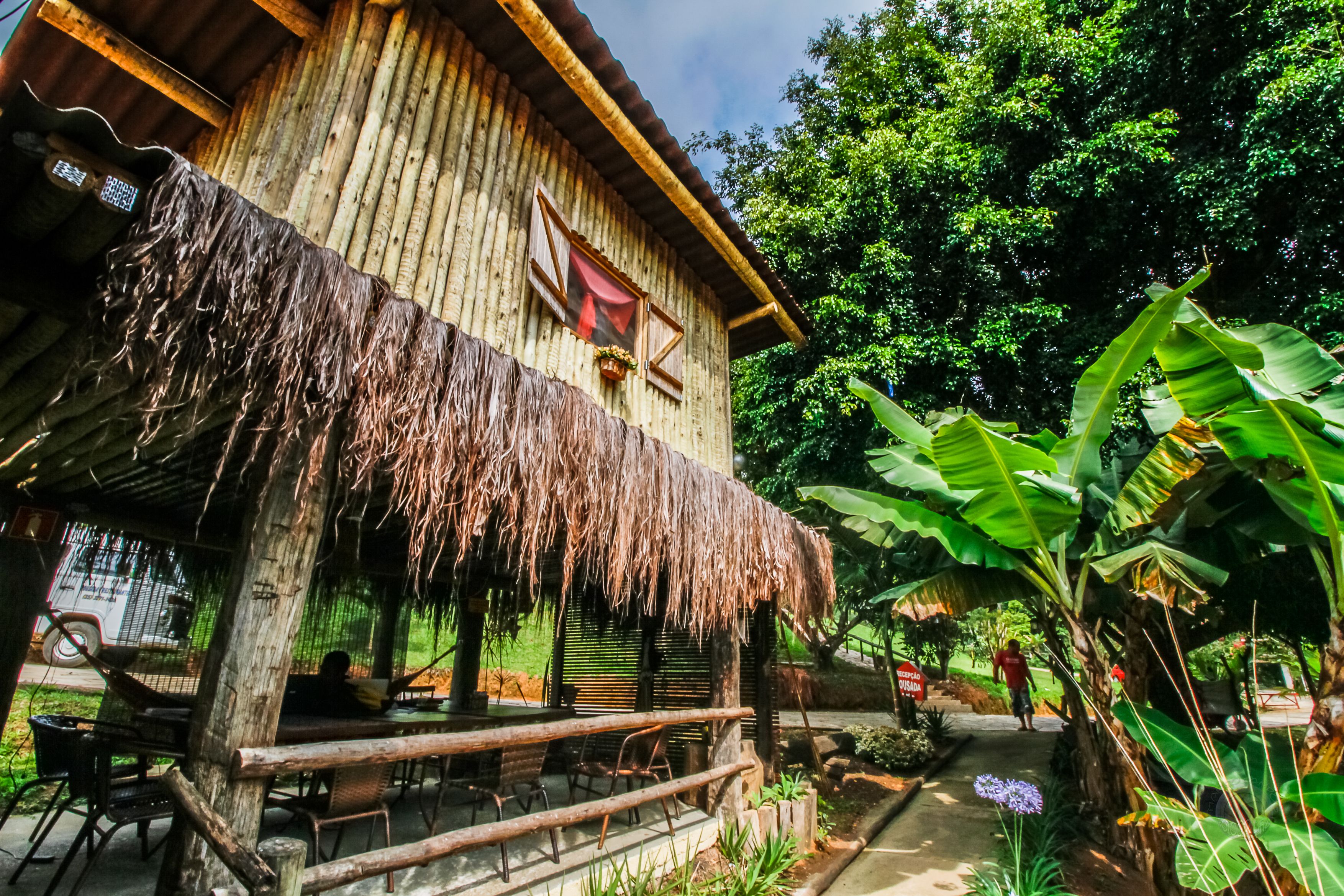A rustic wooden building with a thatched roof, outdoor seating, and lush greenery.
