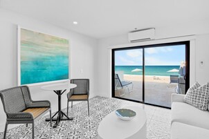 Quarto com vista para a praia, uma pintura grande, uma mesa de jantar, duas cadeiras, um sofá branco e um chão estampado.