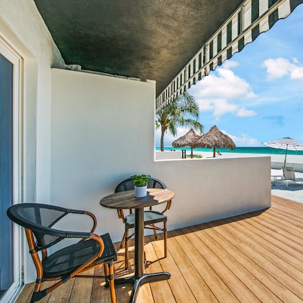 A wooden deck with a table and chairs, a potted plant, and a view of the beach with umbrellas and lounge chairs.