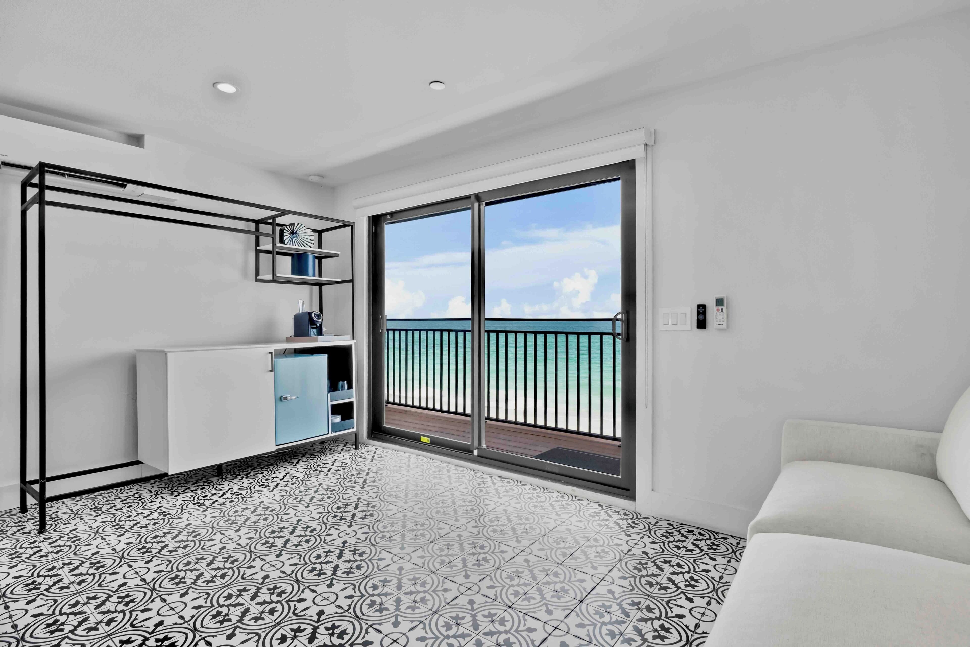 A modern room with a balcony, a white sofa, a kitchen area with a fridge and shelves, and patterned floor tiles.