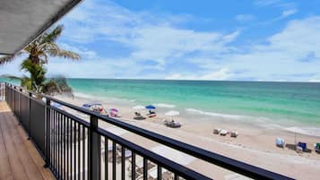 A beach view from a balcony with a wooden floor, palm trees, and beach umbrellas.