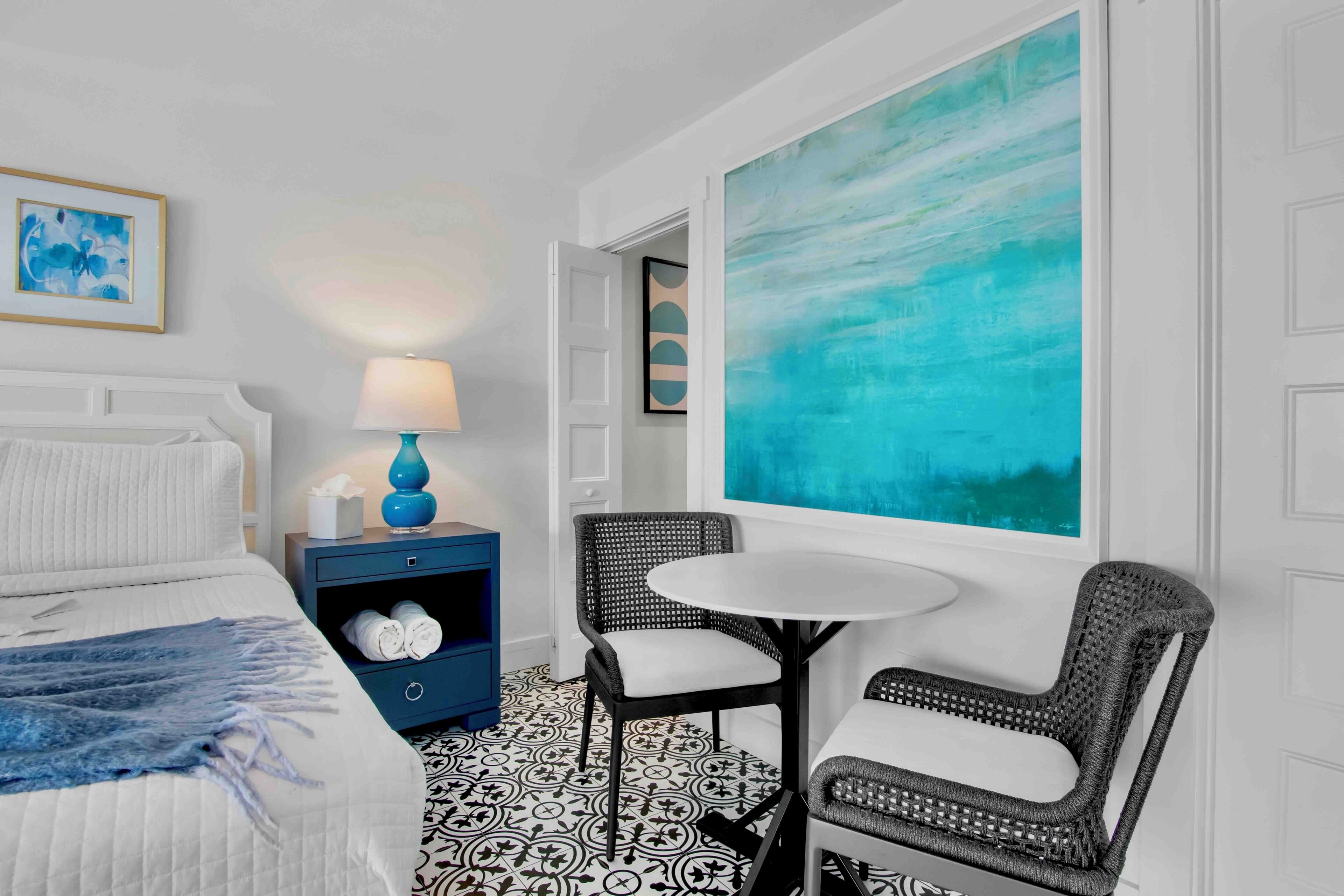 A hotel room with a bed, a nightstand with a lamp, a chair, a table, and a large abstract painting.