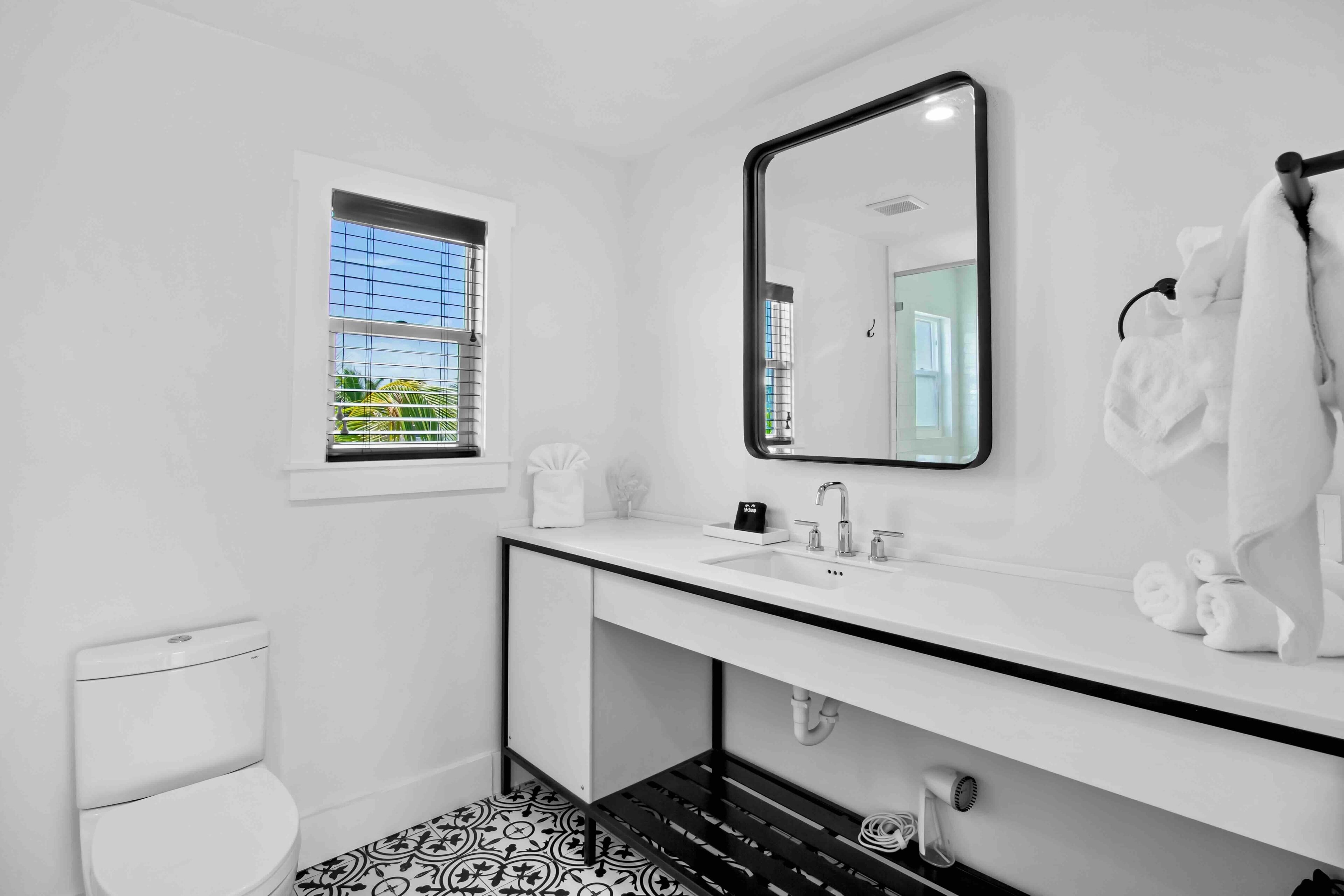 A modern bathroom with a white sink, a large mirror, and a window with blinds.