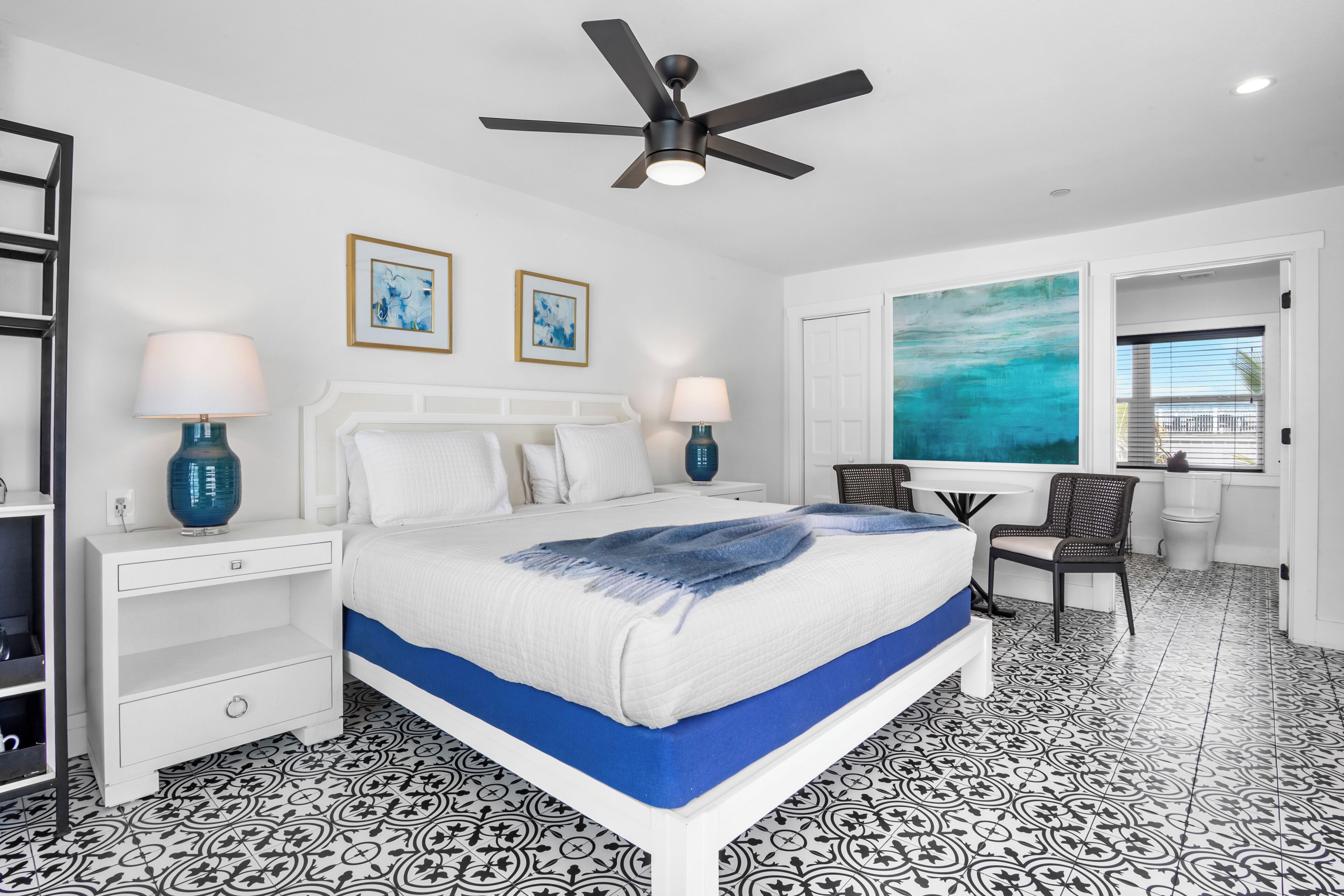 A hotel room with a bed, two bedside tables, a desk, a chair, a ceiling fan, and a large window with a view.