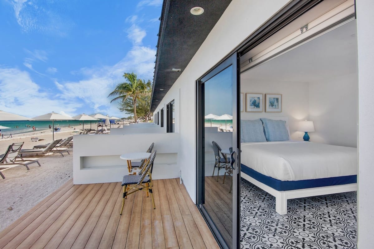 A modern hotel room with a balcony overlooking a beach, a bed with a blue accent, and a view of the ocean.