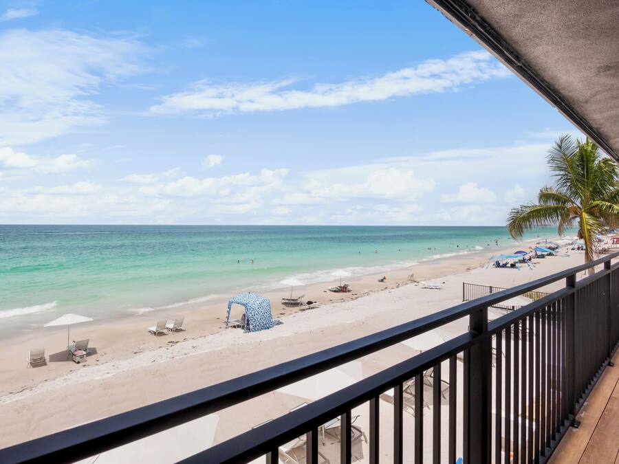 Une vue sur la plage depuis un balcon doté d’une balustrade, avec un palmier et des parasols de plage.