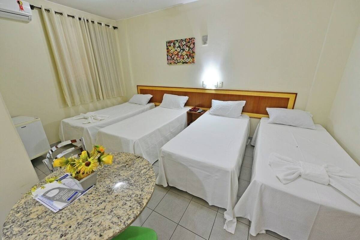 A hotel room with four single beds, a small table with a vase of flowers, a chair, and a wall-mounted air conditioner.