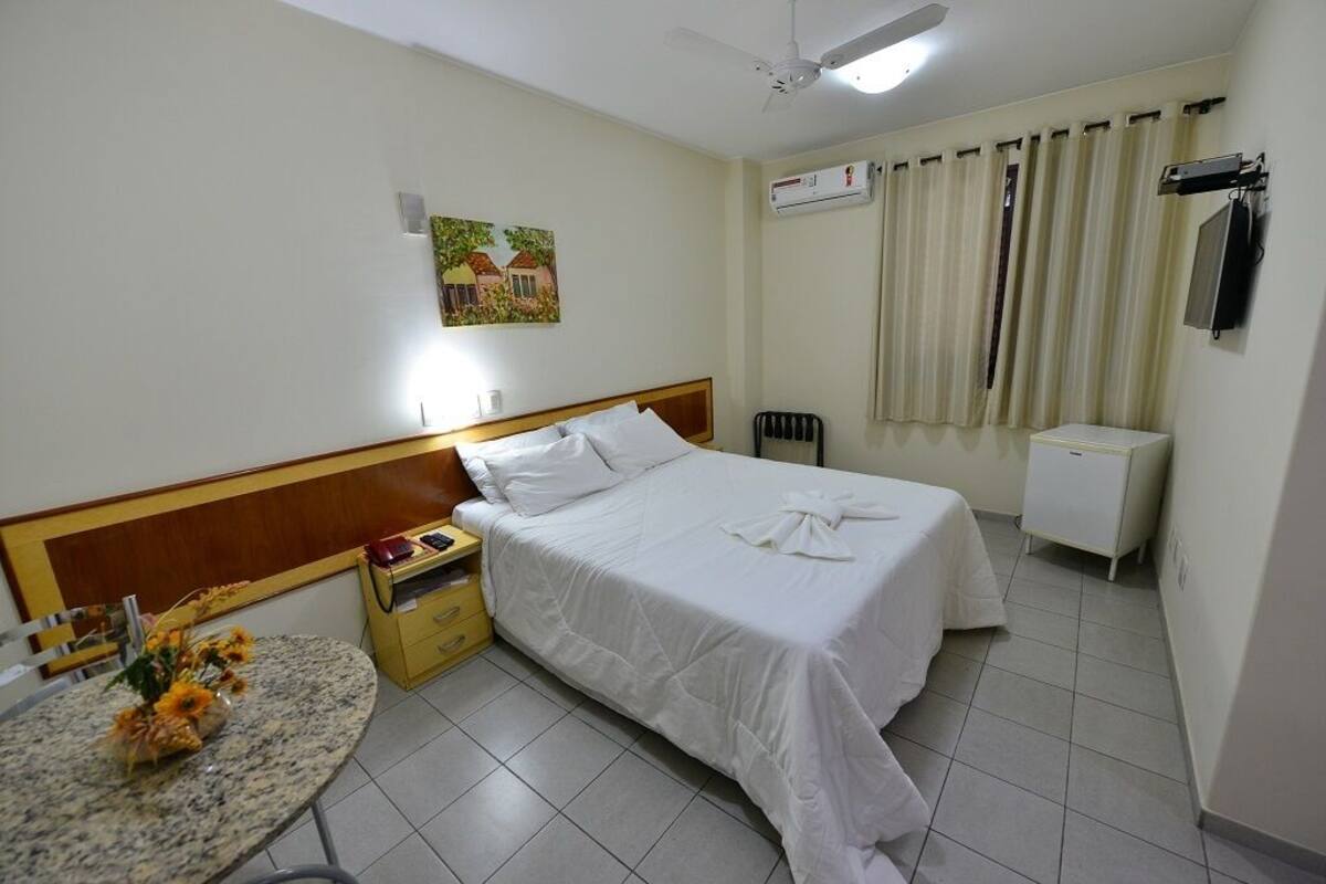 A hotel room with a bed, a bedside table with a lamp, a small table with flowers, a ceiling fan, and an air conditioning unit.