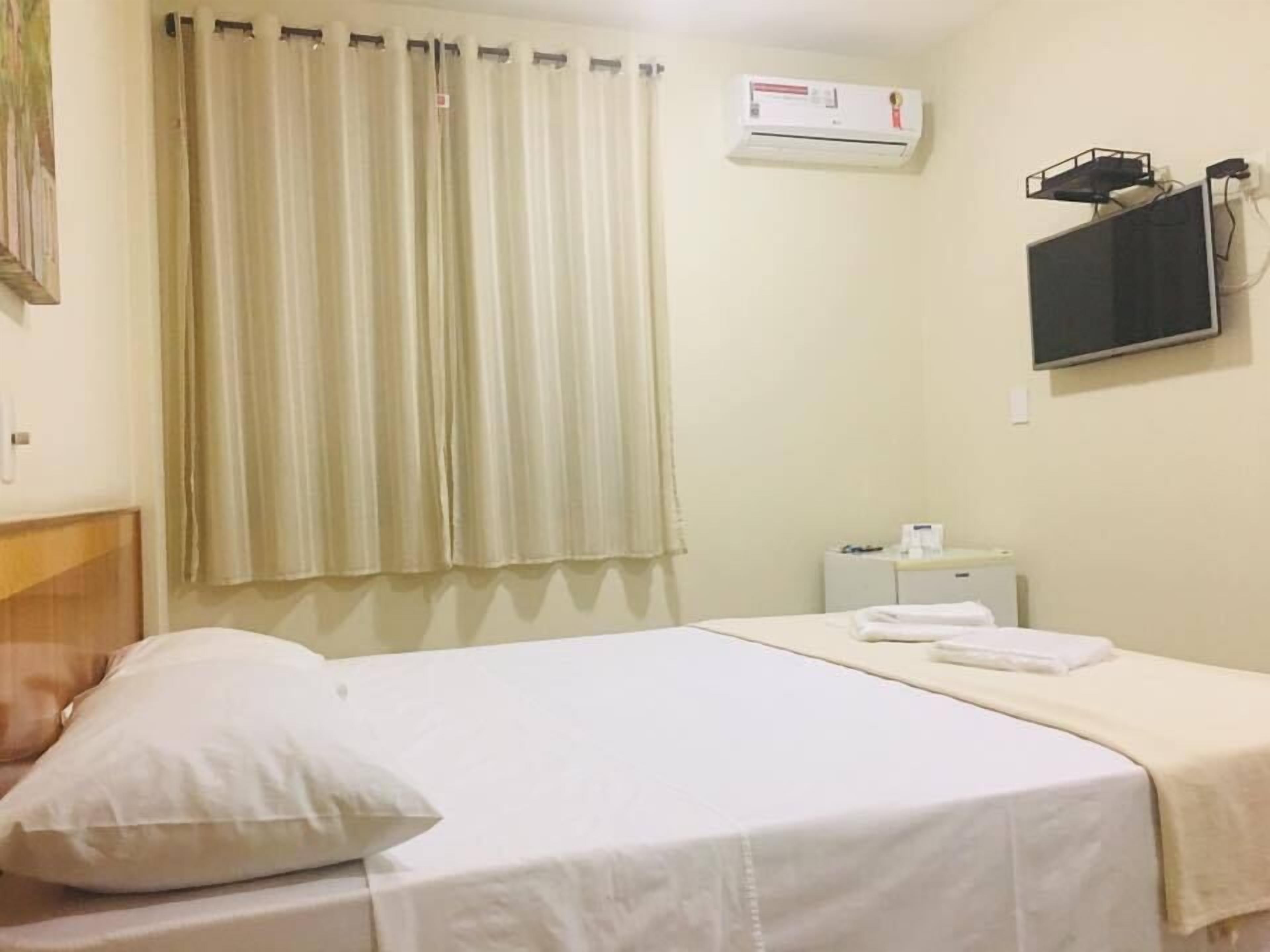 A hotel room with a bed, a pillow, a bedside table with a lamp, a wall-mounted air conditioner, and a television.