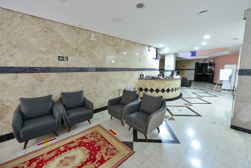 Un hall con mostrador de recepción, tres sillones grises, un tapete estampado y suelo de baldosas con diseños geométricos.