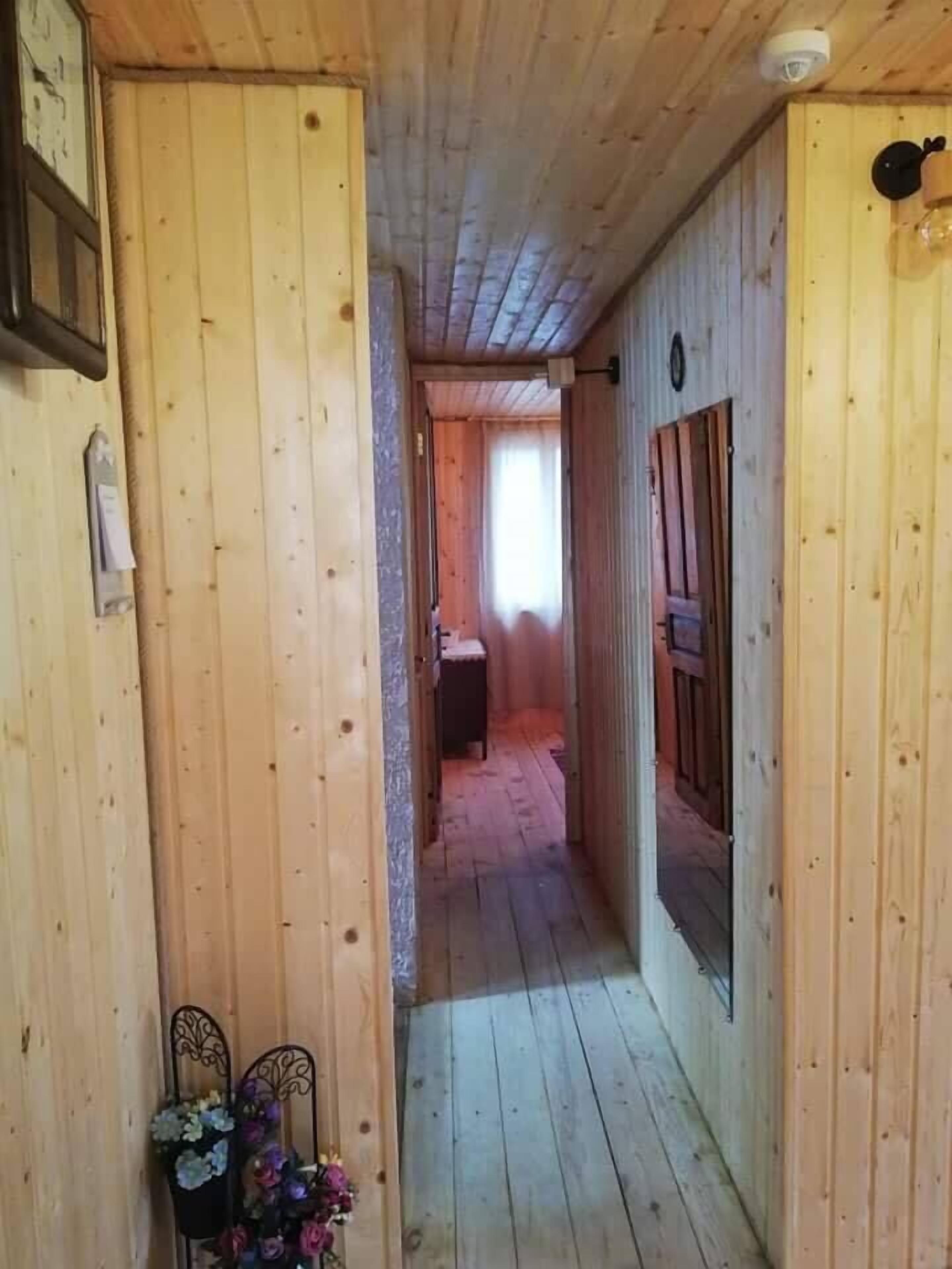 Un estrecho pasillo de madera con una puerta a la derecha, una pequeña mesa con flores a la izquierda y un ventanal al final.