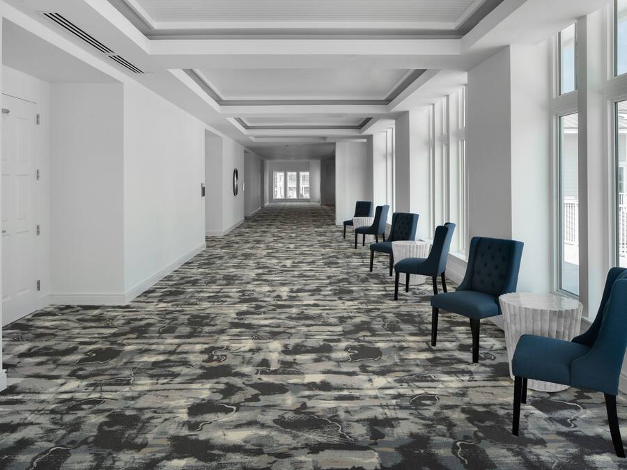 A hotel corridor with a patterned carpet, rows of blue chairs, and large windows.