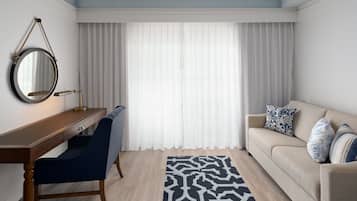 Una habitación moderna con un escritorio de madera, una silla azul, un sofá beige con cojines estampados, un tapete blanco y negro y un ventanal con cortinas translúcidas.