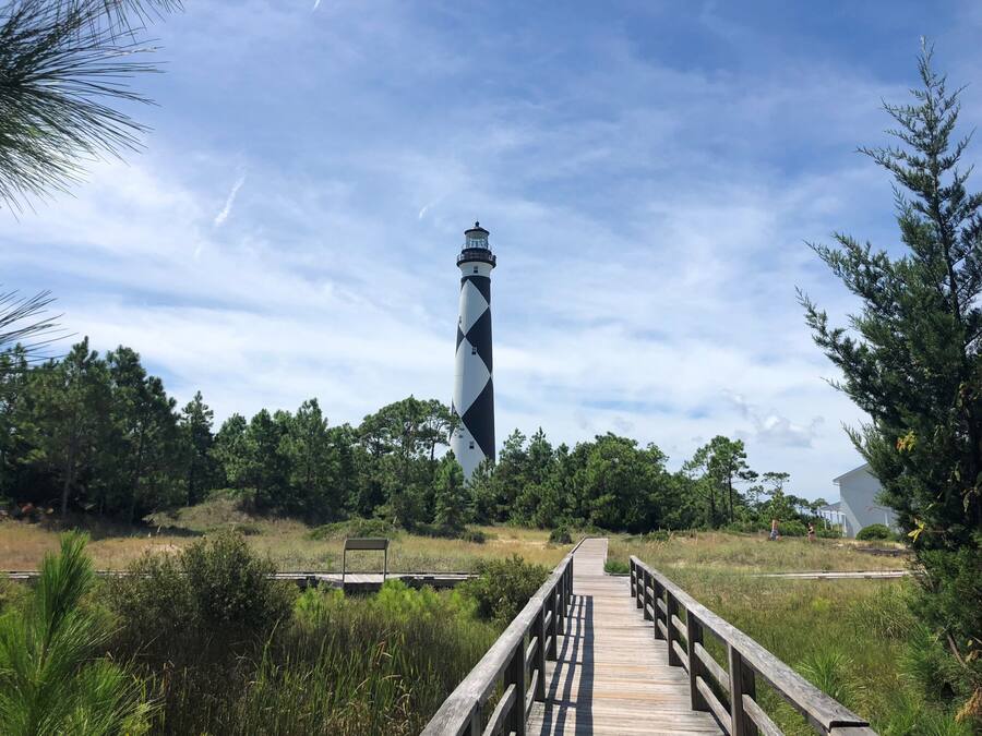 A lighthouse with a distinctive black and white pattern, a wooden walkway leading to it, and surrounding greenery.