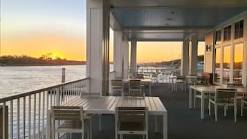 A covered outdoor seating area with tables and chairs, overlooking a body of water during sunset.