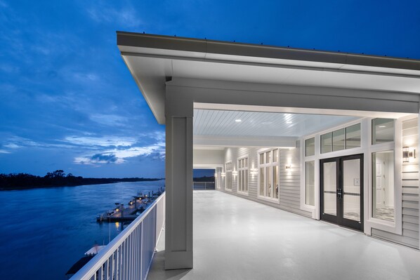 A covered outdoor area with a view of the water, featuring a long corridor with glass doors and windows, and a railing along the side.