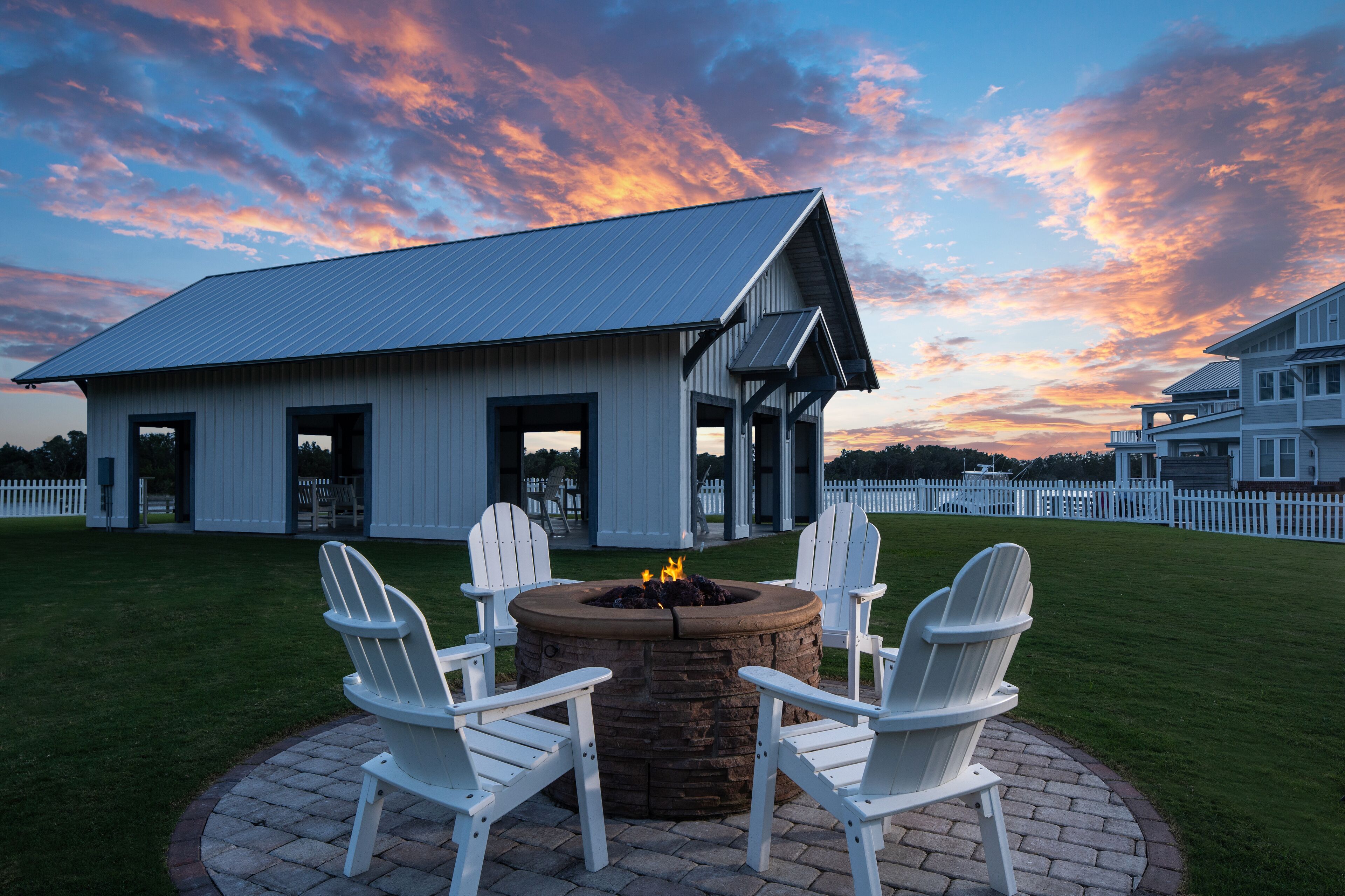A patio with a fire pit and Adirondack chairs, a white building with a metal roof, and a white picket fence in the background.