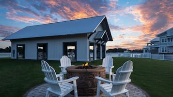A patio with a fire pit and Adirondack chairs, a white building with a metal roof, and a white picket fence in the background.