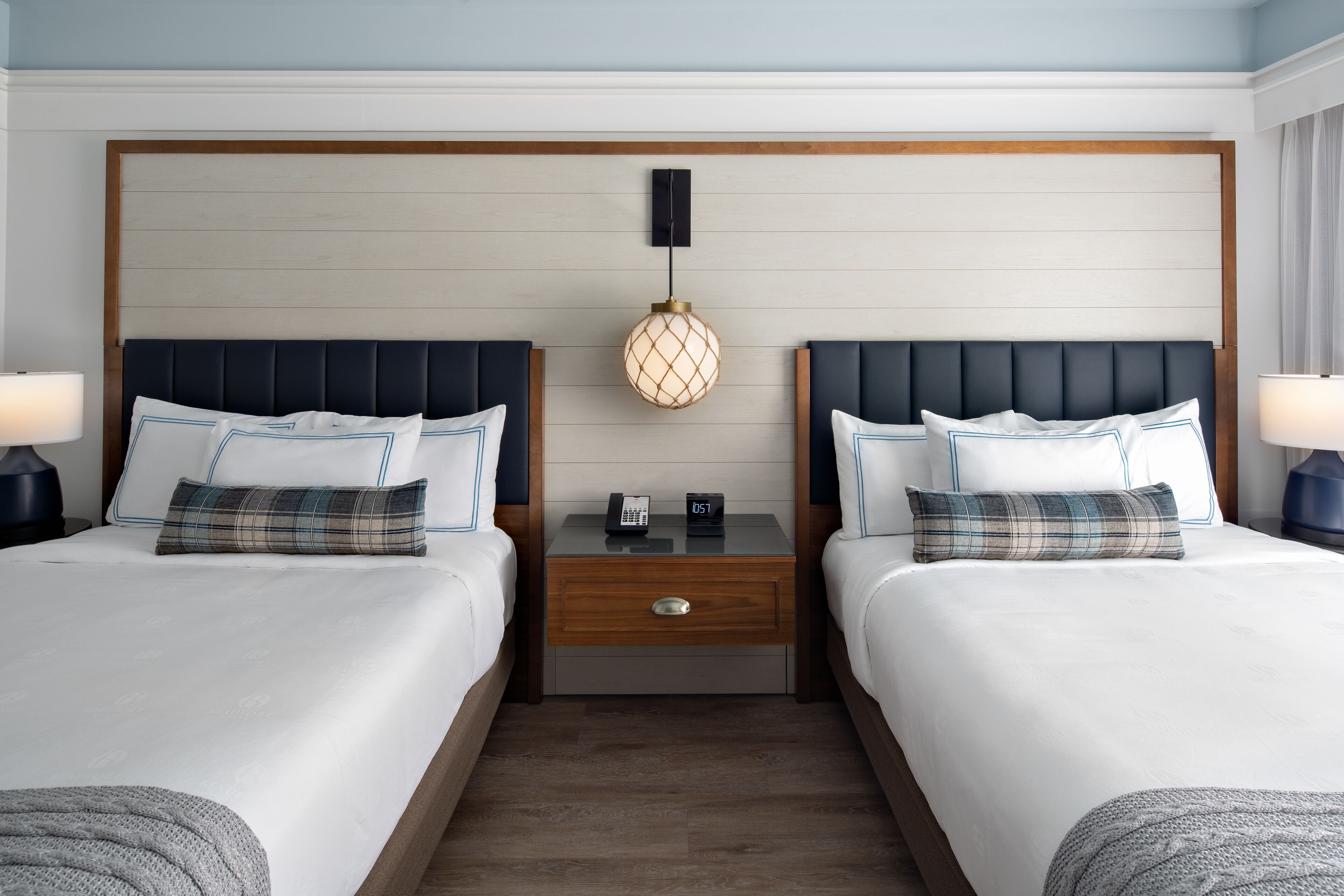 A hotel room with two beds, a wooden headboard, a nightstand with a lamp, and a wall-mounted light fixture.